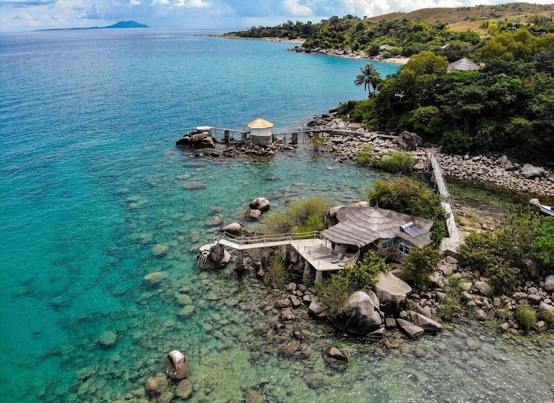 A coastal landscape with clear turquoise waters, a rocky shoreline, a small hut with a thatched roof, and a wooden walkway leading to a dock. 3