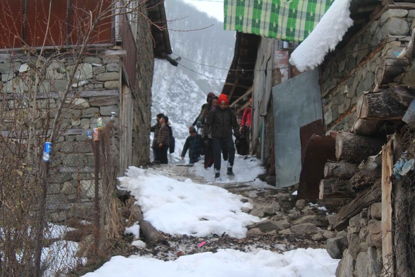 A narrow snow-covered path flanked by stone walls and wooden structures, with people walking in the distance. 2