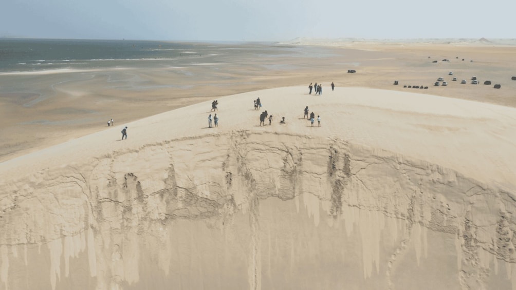 A desert landscape with a group of people and vehicles on a sandy dune, overlooking a beach and ocean. 2