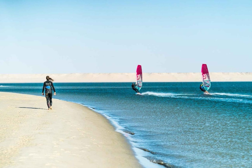 A beach with two windsurfers and a person walking on the sand. 3
