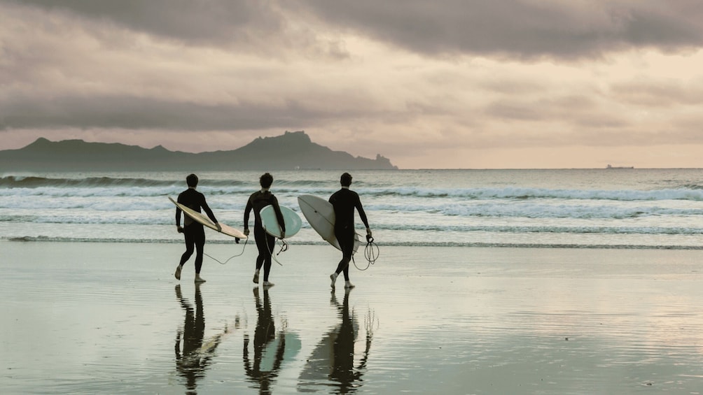 Three surfers walking on a beach with waves in the background and a mountain in the distance. 3