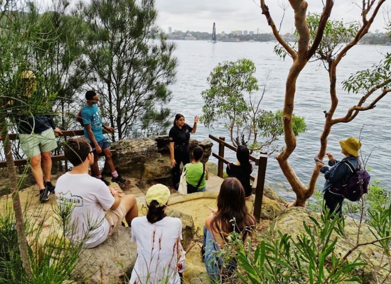 A group of people sitting and standing on rocky terrain by a river, with trees and a bridge in the background. 1
