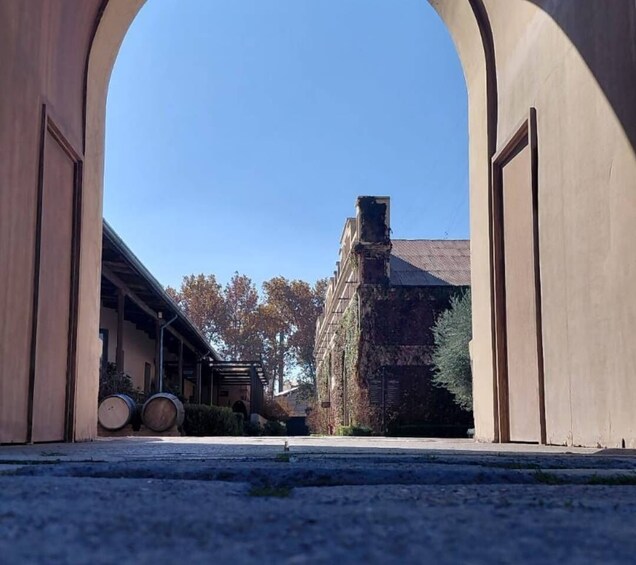 A paved pathway leading to a building with a large arched entrance, barrels, and a tree-lined street. 1