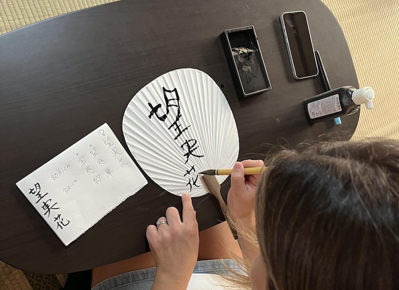 A person writing on a fan with a brush, a piece of paper with handwritten text, and a small box with a brush. 4