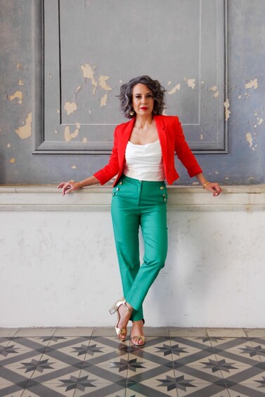 A person standing in front of a wall with peeling paint, wearing a red blazer, white top, green pants, and gold shoes. 3