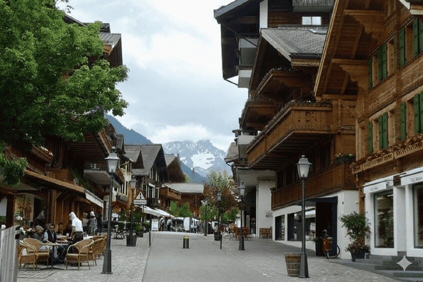 A cobblestone street lined with wooden buildings, a café with patrons, and a mountain in the background. 5