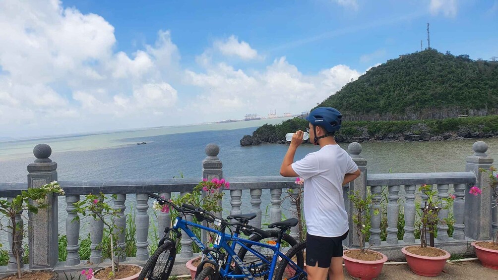 A person drinking water, a blue bicycle, and a scenic view of a body of water with a distant bridge and a hill. 5