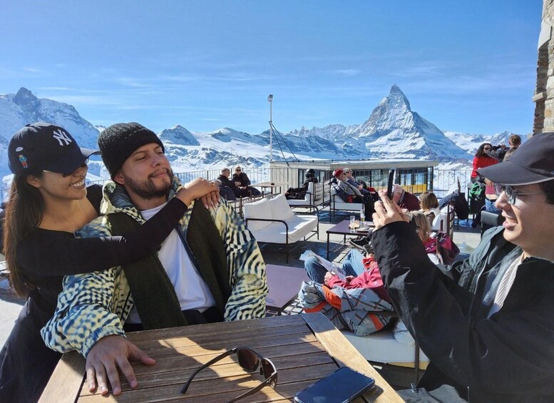 A group of people sitting at a wooden table outdoors with a mountainous landscape in the background. 2
