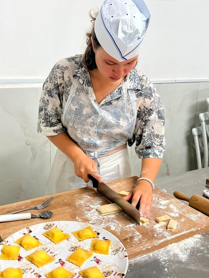 A person cutting dough on a wooden board with a rolling pin and a plate of ravioli. 1