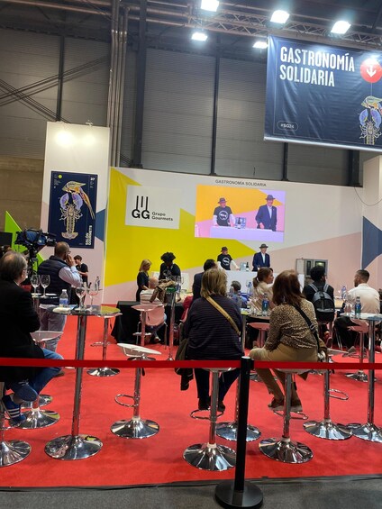 An indoor event with a stage, people seated at tables, and a banner reading 'Gastronomía Solidaria'. 4