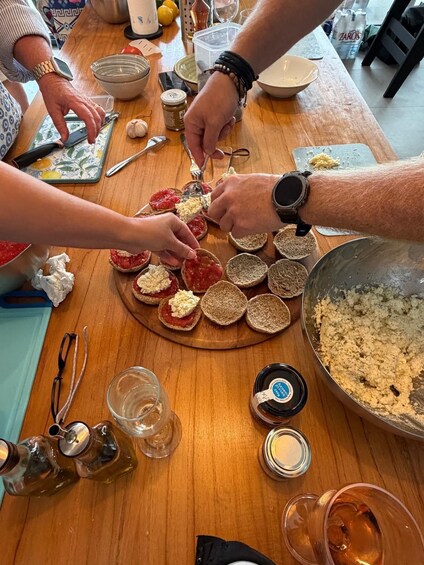 People preparing food on a wooden table with bread, jars, and various kitchen utensils. 2