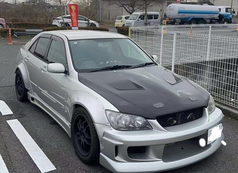 A silver and black car parked in a lot with a white fence and a red sign in the background. 3