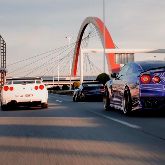 A white sports car, a blue sports car, and a black car driving on a bridge with a red arch. 5