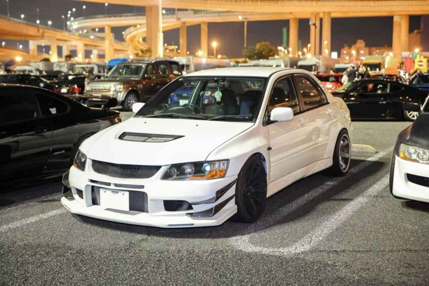 A white car parked in a lot with other vehicles, under a bridge at night. 2