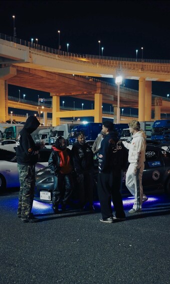 A group of people standing around a car at night under a bridge with streetlights. 4