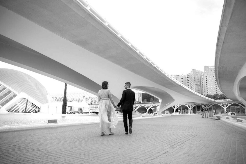 A couple walking under a modern architectural structure with a cityscape in the background. 3