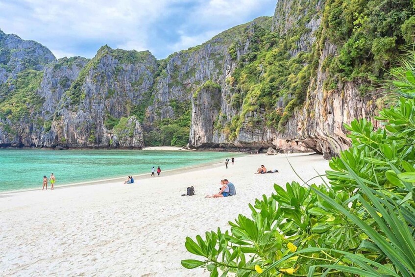 A beach with people sunbathing and walking, surrounded by rocky cliffs and greenery. 2