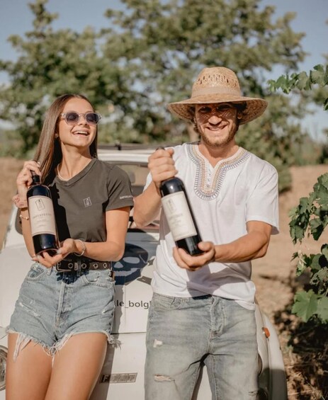 Two people holding wine bottles, standing next to a vehicle in a vineyard. 3