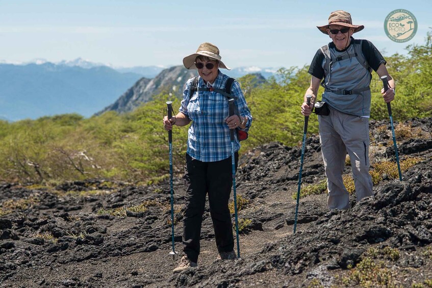 Two people hiking on a volcanic terrain with a mountainous backdrop. 1