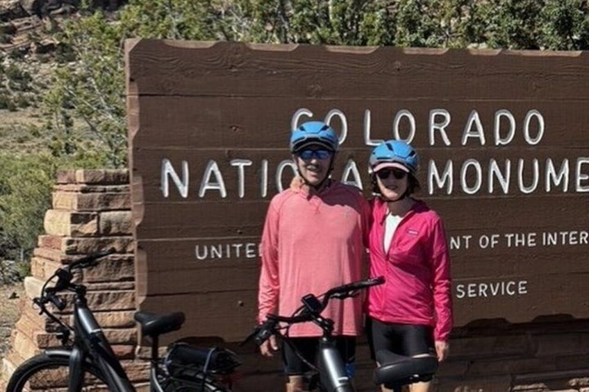 Two people standing in front of a sign that reads 'Colorado National Monument.' 2