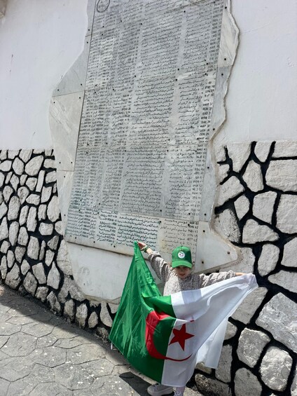 A person holding a flag with a green field and red star, standing in front of a wall with inscriptions. 3