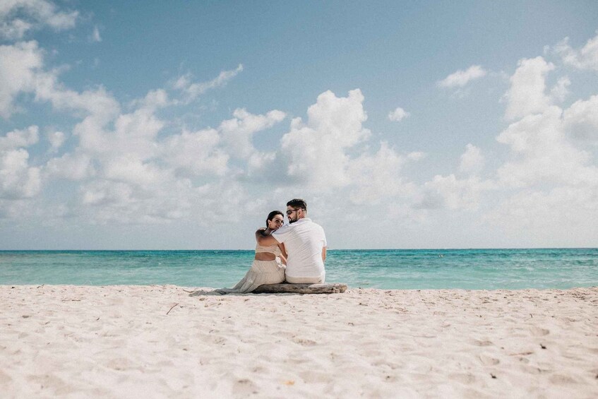 A couple sitting on a wooden log on a sandy beach with turquoise water and a clear sky. 4