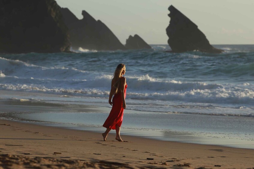 A woman in a red dress walking on a beach with waves and rock formations in the background. 1