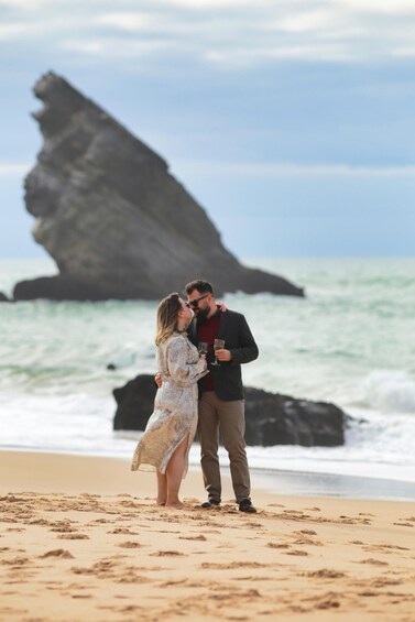A couple embracing on a beach with ocean waves and a large rock formation in the background. 5