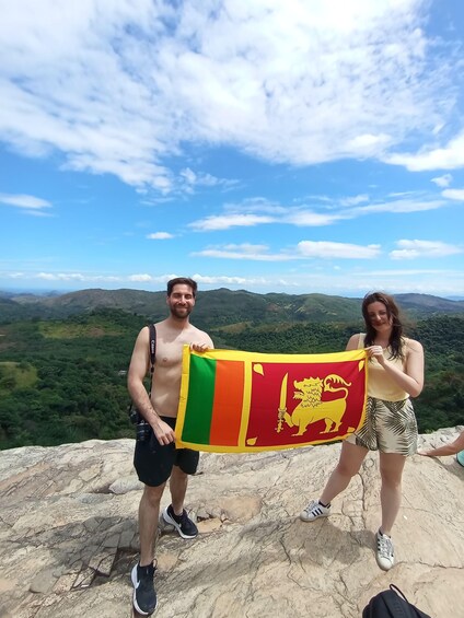 Two people holding a flag on a rocky outcrop with a mountainous landscape in the background. 1
