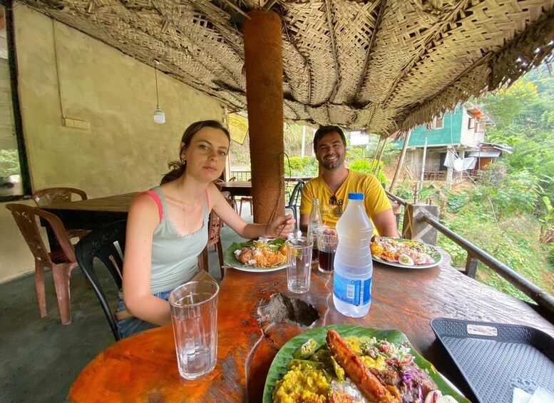 Two people dining at an outdoor table with a woven roof, surrounded by greenery. 2