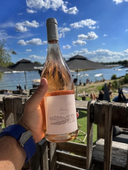 A hand holding a bottle of rosé wine with a label that reads 'Rose 2023' against a backdrop of a lakeside setting with people and umbrellas. 2