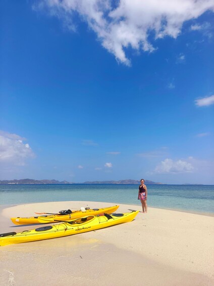 A person standing on a sandy beach with three yellow kayaks in the foreground and a clear blue sky above. 4