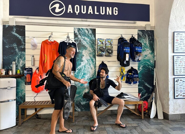 Two people in a diving shop with diving gear and a wetsuit rack. 4