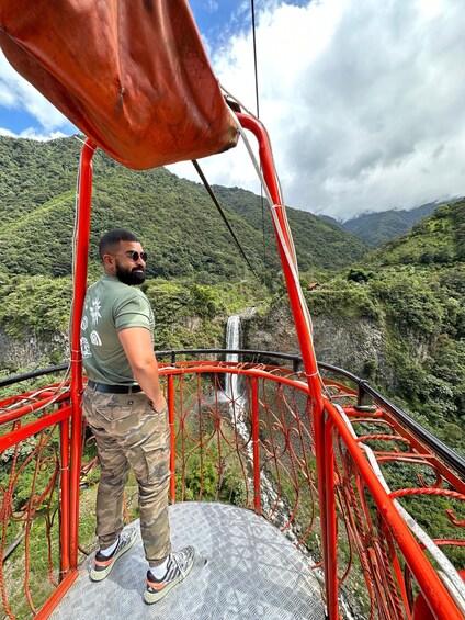 Una persona de pie en una plataforma con barandilla roja, contemplando un paisaje montañoso con cascada. 2