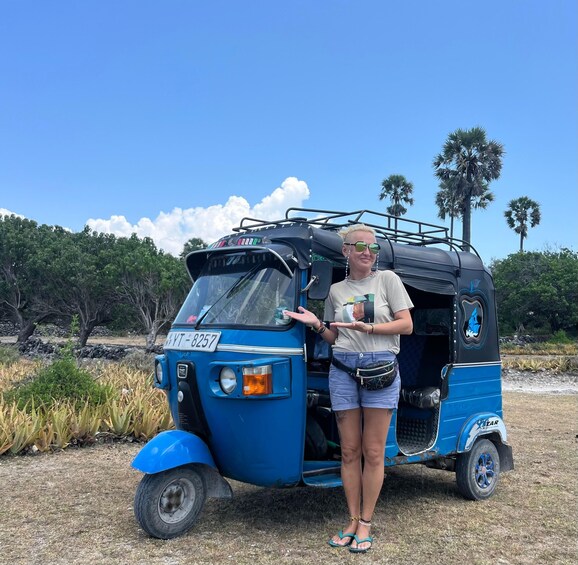A person standing next to a blue tuk-tuk with a license plate 'VT-8257' in a natural setting with trees and dry grass. 1