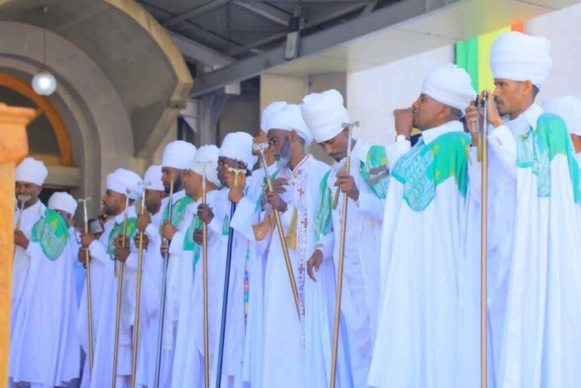 A group of men in white robes with green accents, holding staffs, standing in a line. 1