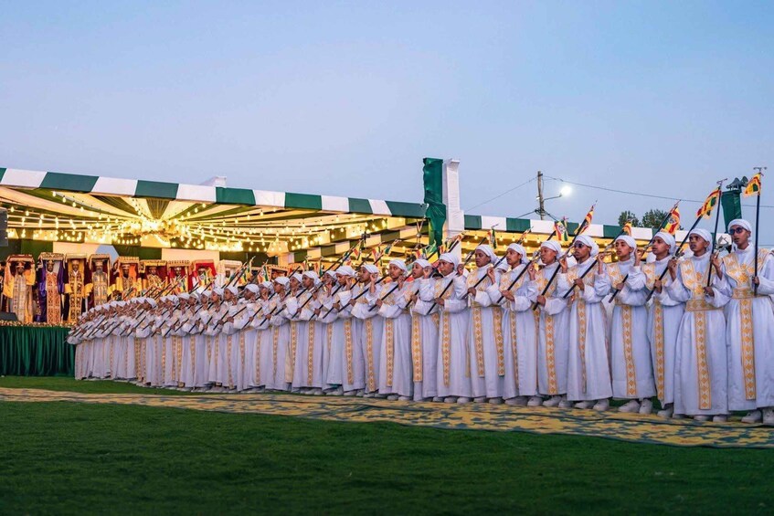 A group of individuals in white robes with gold trim, standing in a line on a grassy field. 2
