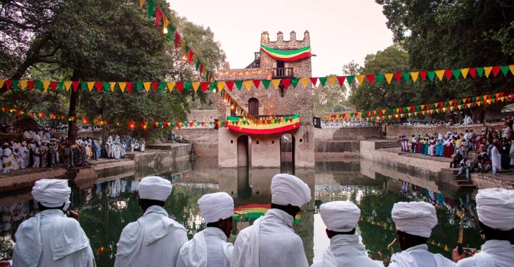 A group of people in white attire gathered around a water body, with a building decorated with colorful flags in the background. 1
