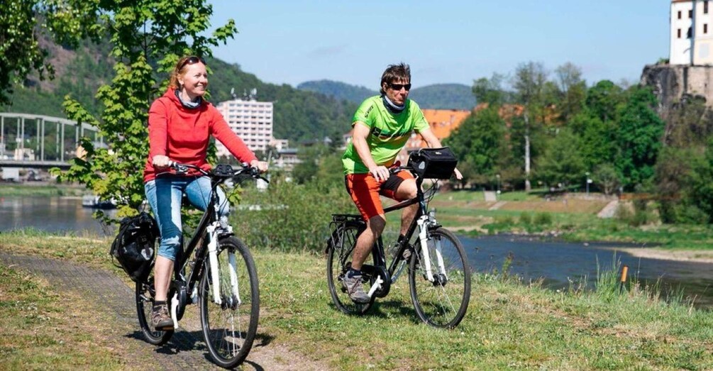 Two cyclists on a path by a river with a bridge and buildings in the background. 3