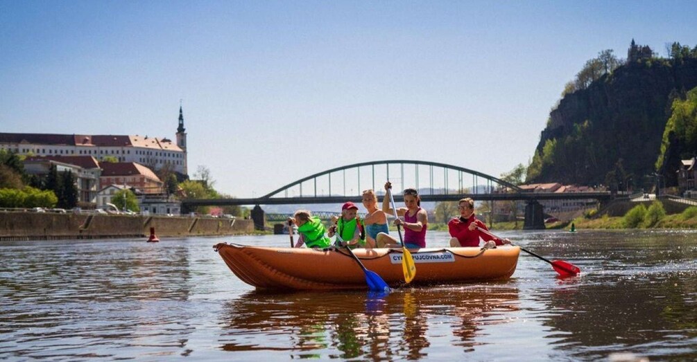 A group of people in a small boat on a river, with a bridge and buildings in the background. 2