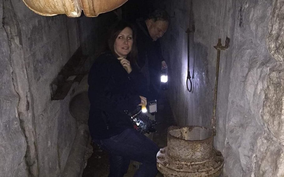 A person in a dimly lit, narrow passageway with rough stone walls, a bucket, and a hook on the wall. 2