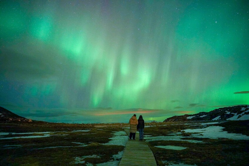 Two people on a wooden boardwalk under a green aurora display. 1