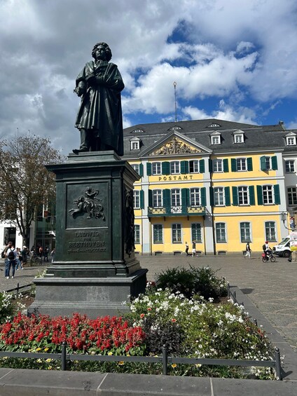 A statue of Ludwig van Beethoven in front of a yellow building with green shutters, surrounded by a flower bed. 1