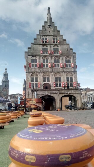 A historic building with a steeply pitched roof and multiple windows, surrounded by a market area with cheese wheels on display. 2