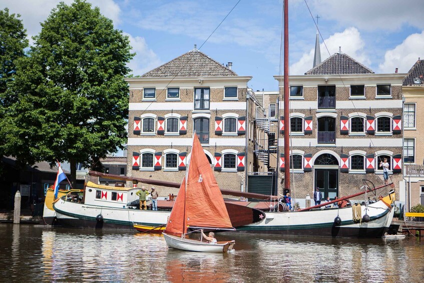A canal with a traditional boat with an orange sail, a tree, and buildings with distinctive window patterns. 1