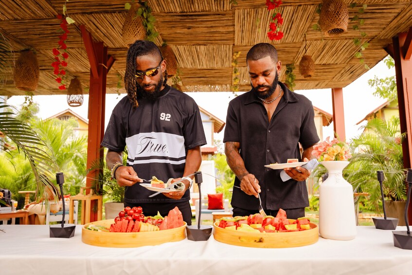 Two men serving food at an outdoor event with tropical plants and a thatched roof. 1