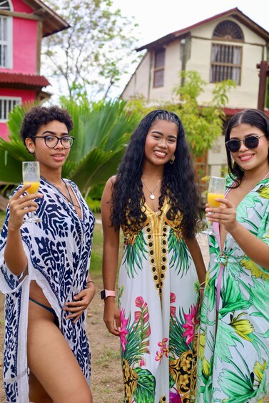 Three people in tropical attire holding drinks, with a house and greenery in the background. 3