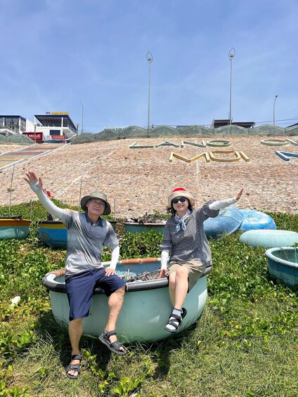 Two people sitting on a boat with arms raised, surrounded by water and greenery, with a large sign in the background. 3