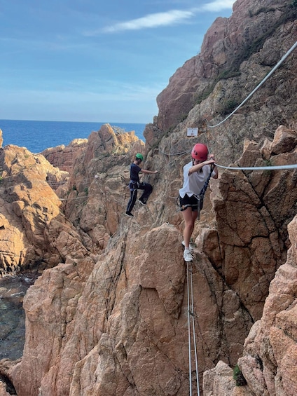 Two individuals rock climbing on a rocky cliff with a rope, overlooking a coastal landscape. 3