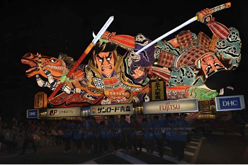 A night scene with a large illuminated display of a samurai warrior, surrounded by spectators. 1